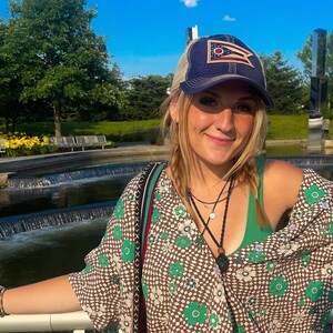 May include: A woman wearing a navy blue and tan trucker hat with a flag design. She is also wearing a patterned top with green and white floral designs. The background includes a water feature and trees.