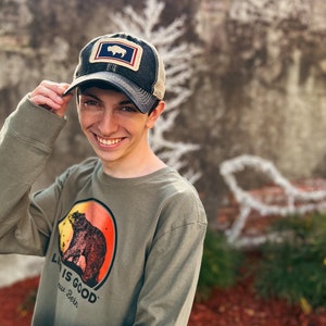 May include: A dark gray trucker hat with a patch featuring a white bison inside a blue and red rectangle. The hat is worn with a long-sleeved olive green shirt with a bear graphic and the text "LIFE IS GOOD New Bern".