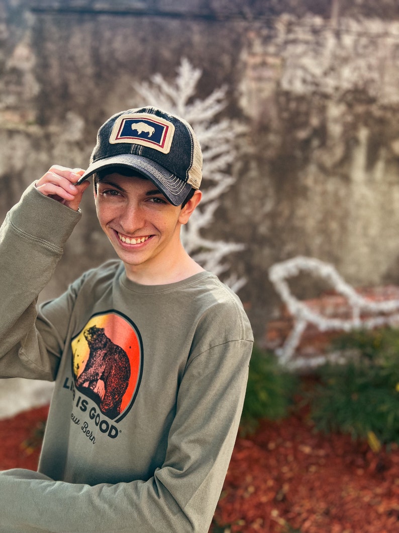 May include: A dark gray trucker hat with a patch featuring a white bison inside a blue and red rectangle. The hat is worn with a long-sleeved olive green shirt with a bear graphic and the text "LIFE IS GOOD New Bern".