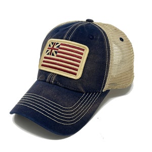 May include: A navy blue trucker hat with a beige mesh back and a curved brim. The hat features a rectangular patch with a flag design, combining elements of the American and British flags. The stitching is in a contrasting beige color.