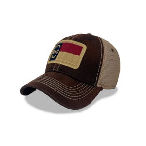 May include: A brown baseball cap with a tan mesh back. The front features a patch with a flag design in red, tan, and blue. The brim has white stitching.