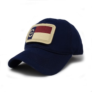 May include: A navy blue baseball cap with a rectangular patch featuring a flag design. The flag has a red and white stripe, a blue circle with stars, and a tan border. The cap has a curved brim.