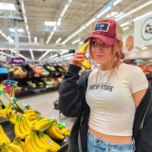 May include: A young person wearing a white shirt with the text "Brooklyn New York" on it, a maroon and beige baseball cap with a palm tree design, and blue jeans. The person is holding a banana to their ear like a phone. The person is standing in a grocery store aisle with bananas in the background.