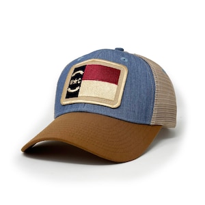 May include: A blue and brown trucker hat with a patch of the North Carolina state flag. The flag is red, white, and gold with a star and the letters "N*C" in a circle.