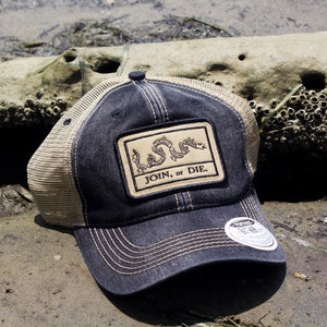 May include: A navy blue and tan trucker hat with a patch featuring a coiled snake and the text "JOIN, OR DIE." The hat has a curved brim and a mesh back. The hat is sitting on a sandy surface.