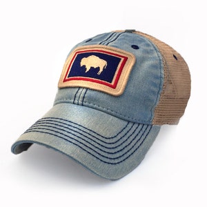 May include: A blue denim baseball cap with a tan mesh back. The cap features a rectangular patch with a white bison silhouette on a blue background, framed by red and tan borders. The cap has a curved brim and is suitable for outdoor activities.