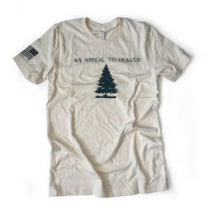May include: Cream-colored t-shirt with the text "AN APPEAL TO HEAVEN" above a dark green pine tree graphic. The left sleeve features a small American flag design. The shirt is made of a soft, lightweight material.