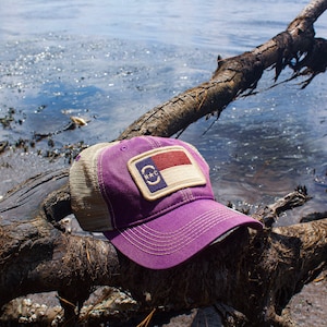 May include: A purple baseball cap with a mesh back and a patch featuring the North Carolina state flag. The hat is resting on a weathered log near water.