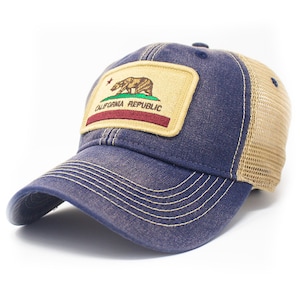 May include: A blue and tan trucker hat with a patch featuring the California Republic flag. The flag has a brown bear, a red stripe, and a green stripe. The text "CALIFORNIA REPUBLIC" is embroidered on the patch.