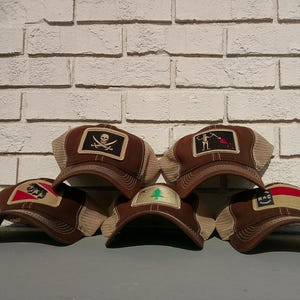 May include: A collection of brown trucker hats with various embroidered patches. Patches include a skull and crossbones, a skeleton with a red flag, a bear, a pine tree, and a flag. The hats have a mesh back and a curved brim.