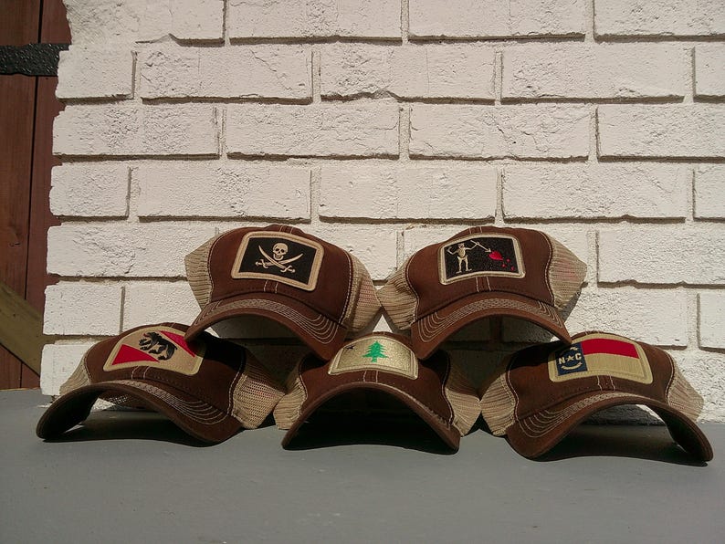 May include: A collection of brown trucker hats with various embroidered patches. Patches include a skull and crossbones, a skeleton with a red flag, a bear, a pine tree, and a flag. The hats have a mesh back and a curved brim.