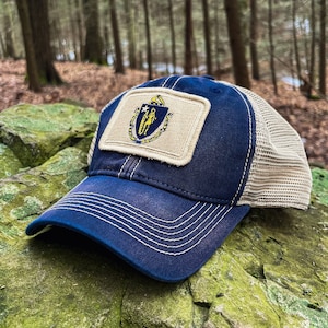 May include: A navy blue and beige trucker hat with a patch featuring the state seal of Massachusetts. The patch is beige with a gold outline and features a gold shield with a blue and white design.