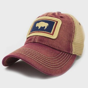 May include: A maroon baseball cap with a mesh back and a curved brim. The front features a rectangular patch with a gold bison silhouette on a navy blue background, framed by a gold border. The cap has a distressed, vintage look.