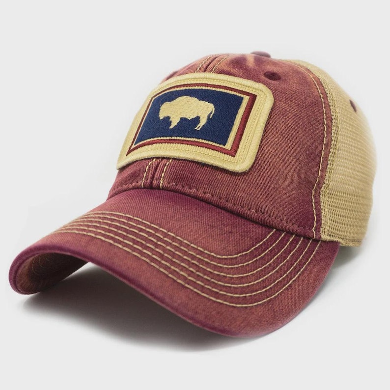 May include: A maroon baseball cap with a mesh back and a curved brim. The front features a rectangular patch with a gold bison silhouette on a navy blue background, framed by a gold border. The cap has a distressed, vintage look.