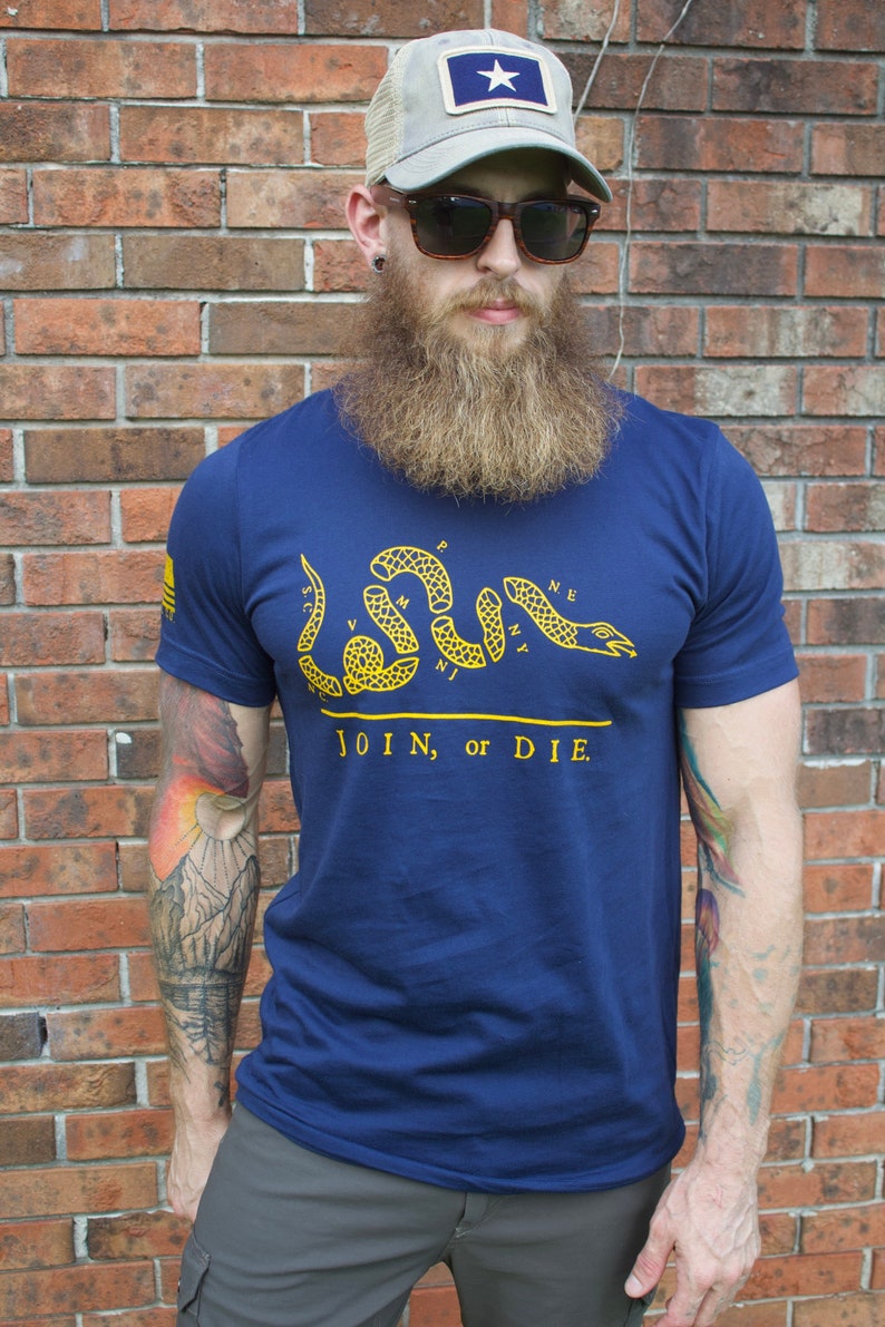 K&ouml;nnte beinhalten: Marineblaues T-Shirt mit gelber Schlangengrafik und dem Text "JOIN, or DIE". Der Mann tr&auml;gt eine graue Trucker-M&uuml;tze mit einem blauen Aufn&auml;her mit einem wei&szlig;en Stern. Der Mann tr&auml;gt eine Sonnenbrille.