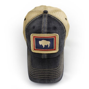 May include: A black and tan trucker hat with a patch featuring a bison silhouette on a blue background, framed by red and tan borders. The hat has a mesh back and a curved brim, with visible stitching details.