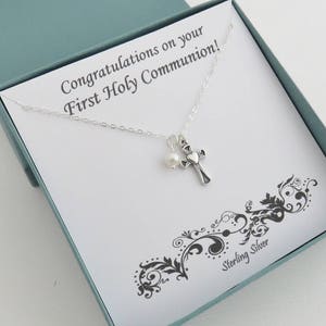 May include: A white card with the text "Congratulations on your First Holy Communion!" and a silver necklace with a cross charm and a pearl bead. The card has a black and white floral design and the text "Sterling Silver" at the bottom.