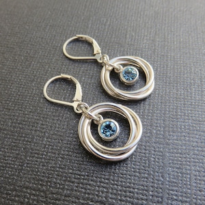 May include: A pair of silver earrings with a blue crystal in the center of each earring. The earrings have three interlocking circles.