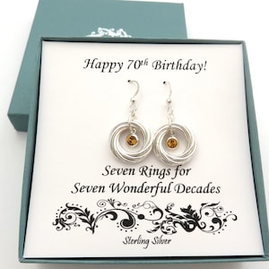 May include: A pair of silver earrings with a circular design, each featuring a small, amber-colored gemstone. The earrings are presented in a teal gift box with the text "Happy 70th Birthday!" and "Seven Rings for Seven Wonderful Decades."