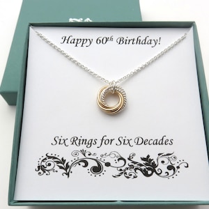 May include: A silver necklace with a gold and silver intertwined ring pendant in a green gift box. The box has a white card inside with the text "Happy 60th Birthday!" and "Six Rings for Six Decades".