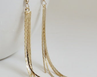 May include: A pair of gold-colored dangle earrings. Each earring features a small ball at the top, a textured rectangular section, and multiple strands of gold chains that cascade downwards. The earrings are set against a white background.