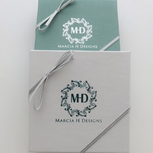 May include: Two gift boxes with silver ribbon bows. The top box is light blue with a white logo that says "MH" inside a decorative circle and "Marcia H Designs" below. The bottom box is white with a dark blue logo that says "MH" inside a decorative circle and "Marcia H Designs" below.