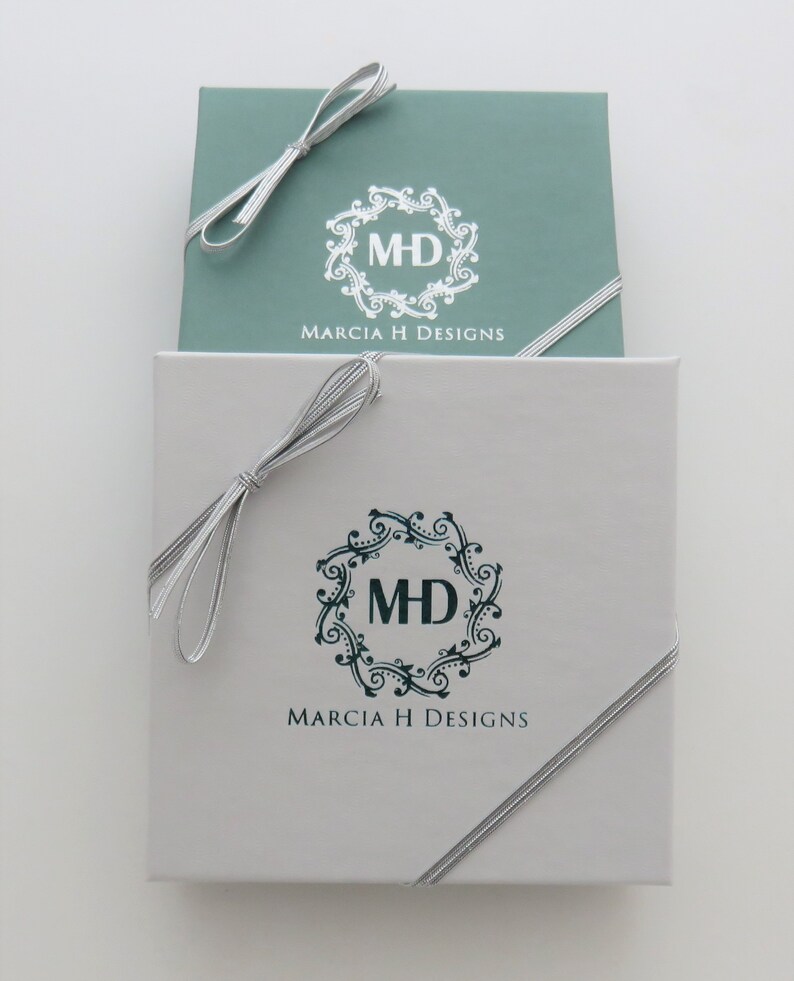 May include: Two gift boxes with silver ribbon bows. The top box is light blue with a white logo that says "MH" inside a decorative circle and "Marcia H Designs" below. The bottom box is white with a dark blue logo that says "MH" inside a decorative circle and "Marcia H Designs" below.