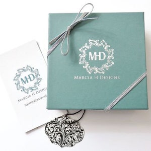 May include: A teal gift box with a silver ribbon and a white logo that says "Marcia H Designs" and "MHD" in a decorative circle. The box is sitting on a white surface.