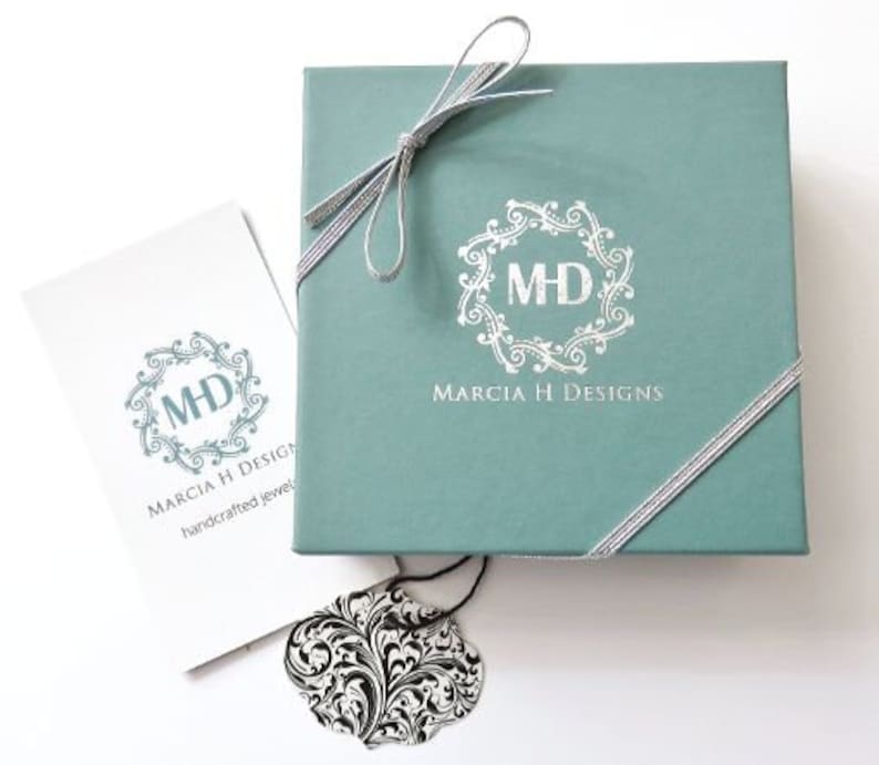 May include: A teal gift box with a silver ribbon and a white logo that says "Marcia H Designs" and "MHD" in a decorative circle. The box is sitting on a white surface.