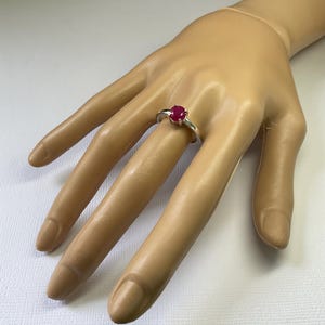 May include: A silver ring featuring a single oval-cut vibrant red gemstone set in a four-prong mounting. The band is smooth and polished, showcasing a simple design that highlights the deep red colour of the central stone on a plain surface.
