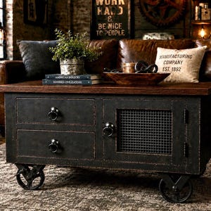 May include: A rectangular coffee table with a dark brown wooden top and a distressed black metal base. The base features two stacked drawers with round ring pulls and a side cabinet door with a black metal mesh panel, all supported by four black wheels.