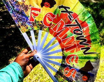 May include: A colorful hand fan with a blue handle and a yellow, green, and red design. The fan features the text "I am from" and "Hometown" in red and black lettering.