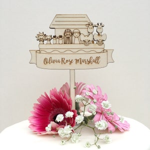 May include: Wooden Noah's Ark cake topper with a banner that reads "Olivia Rose Marshall". The ark is decorated with animals and people. The topper is on a white cake with pink flowers.