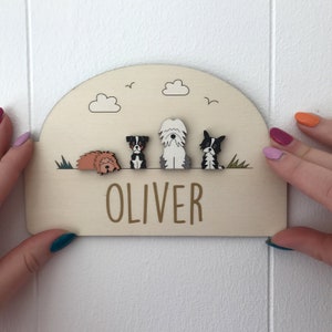 May include: A wooden sign with the name "OLIVER" in brown lettering. The sign features a row of four dog illustrations, grass, clouds, and birds. The sign is held by two hands with painted nails.