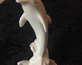 May include: A white porcelain dolphin figurine with gold accents. The dolphin is leaping out of the water, with its body arched and mouth open. The base features wave-like details with gold trim. A decorative nautical accent.