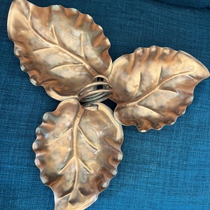 May include: A copper-colored, three-leaf decorative dish. The leaves are detailed with veining and connected by a central, coiled stem. The dish is likely made of metal and has a shiny finish. The overall design is reminiscent of nature.
