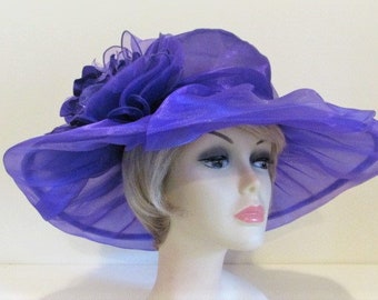 Pode incluir: Um chapéu roxo de aba larga com uma grande flor franjada de um lado. O chapéu é feito de um tecido leve e transparente.