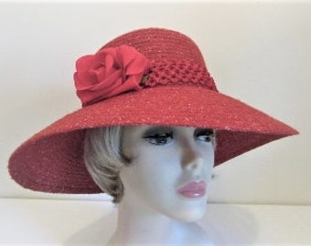 Pode incluir: Um chapéu de sol de aba larga vermelho com uma grande rosa de tecido vermelho no lado. O chapéu tem uma guarnição trançada em torno da coroa.