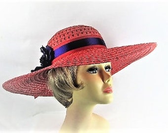 Pode incluir: Chapéu de sol coral com aba larga e design tecido. Apresenta uma fita azul marinho e preta e uma flor preta. O chapéu é exibido em uma cabeça de manequim, mostrando seu design e forma.