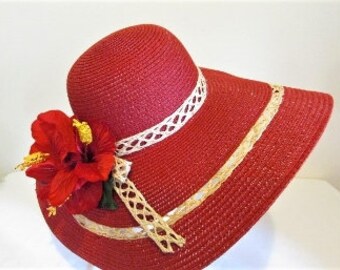 Pode incluir: Um chapéu de sol de palha vermelho com aba larga e guarnição de renda branca. Uma grande flor vermelha está presa ao lado do chapéu.