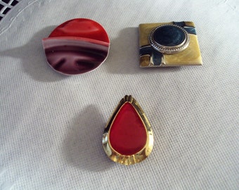 May include: Three decorative brooches with gold accents. One is a red circle with a white stripe, another is a teardrop shape with a red center, and the third is a square with a blue circle and gold and black stripes.