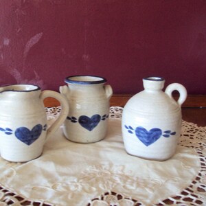 May include: Three small ceramic jugs with blue and white heart designs. The jugs are on a white crocheted doily.