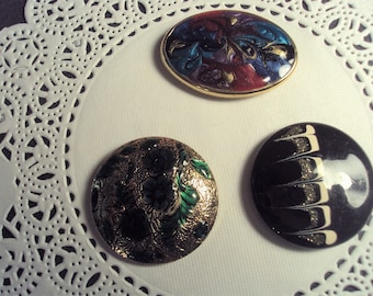 May include: Three decorative buttons with different designs. One button is oval shaped with a gold rim and features a swirling pattern in shades of red, blue, and gold. Another button is round and features a black and white swirl design. The third button is round and features a gold rim with a floral pattern in shades of green and gold.