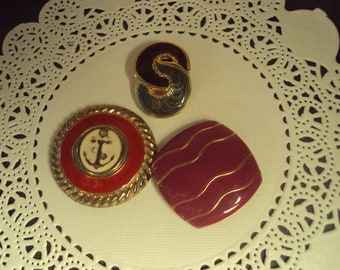 May include: Three decorative brooches with gold trim. One brooch is red with a white anchor, one is burgundy with gold stripes, and one is black and green with a swirl design.