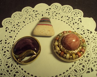 May include: Three decorative brooches with different designs. One is a round brooch with a gold frame and a pink and white swirl design. Another is a round brooch with a purple swirl design and a gold frame. The third is a triangular brooch with a white and brown swirl design.