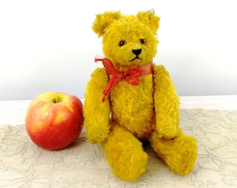 Puede incluir: Un oso de peluche vintage amarillo con una cinta roja alrededor del cuello. El oso está sentado sobre un mantel de encaje blanco.
