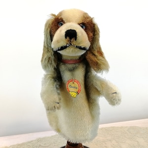 May include: A vintage hand puppet of a brown and white dog with a pink collar. The puppet has a tag that says "Steiff Original  Cocker".