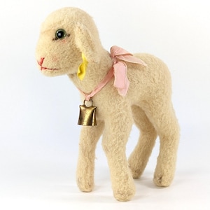 May include: A vintage-style lamb figurine with a cream-colored woolly texture. It features a pink ribbon bow, a yellow ear accent, and a small brass bell around its neck. The lamb has black button eyes and red accents on its face.