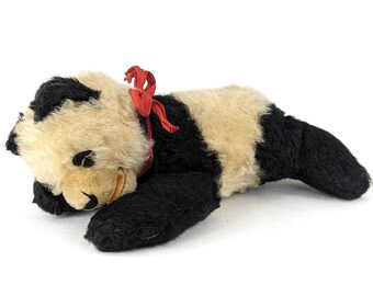 Puede incluir: Un juguete de peluche vintage con forma de oso panda con pelaje negro y crema. El panda tiene una cinta roja alrededor del cuello y está tumbado. Un juguete clásico para niños.