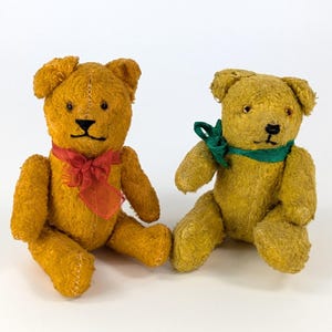 Op de afbeelding: Twee vintage teddyberen met goudkleurige vacht. Eén beer draagt een rood lint, de ander een groene sjaal. Beide hebben zwart gestikte kenmerken en zitten. Een klassiek speelgoed.